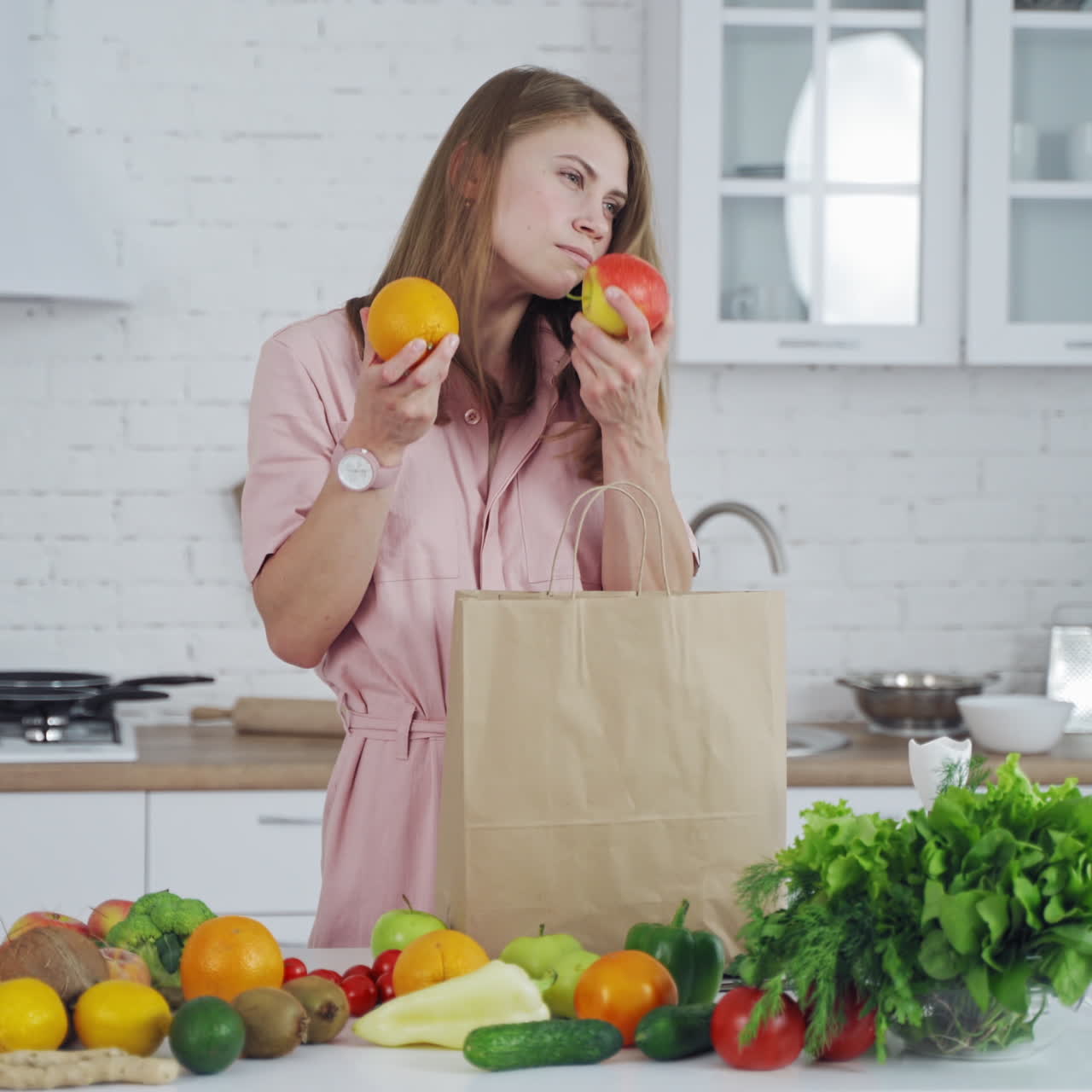 Pretty woman takes fresh fruit from paper bag. Young model is sniffing organic fruit on the light kitchen background. Many healthy food on a table for losing weight.