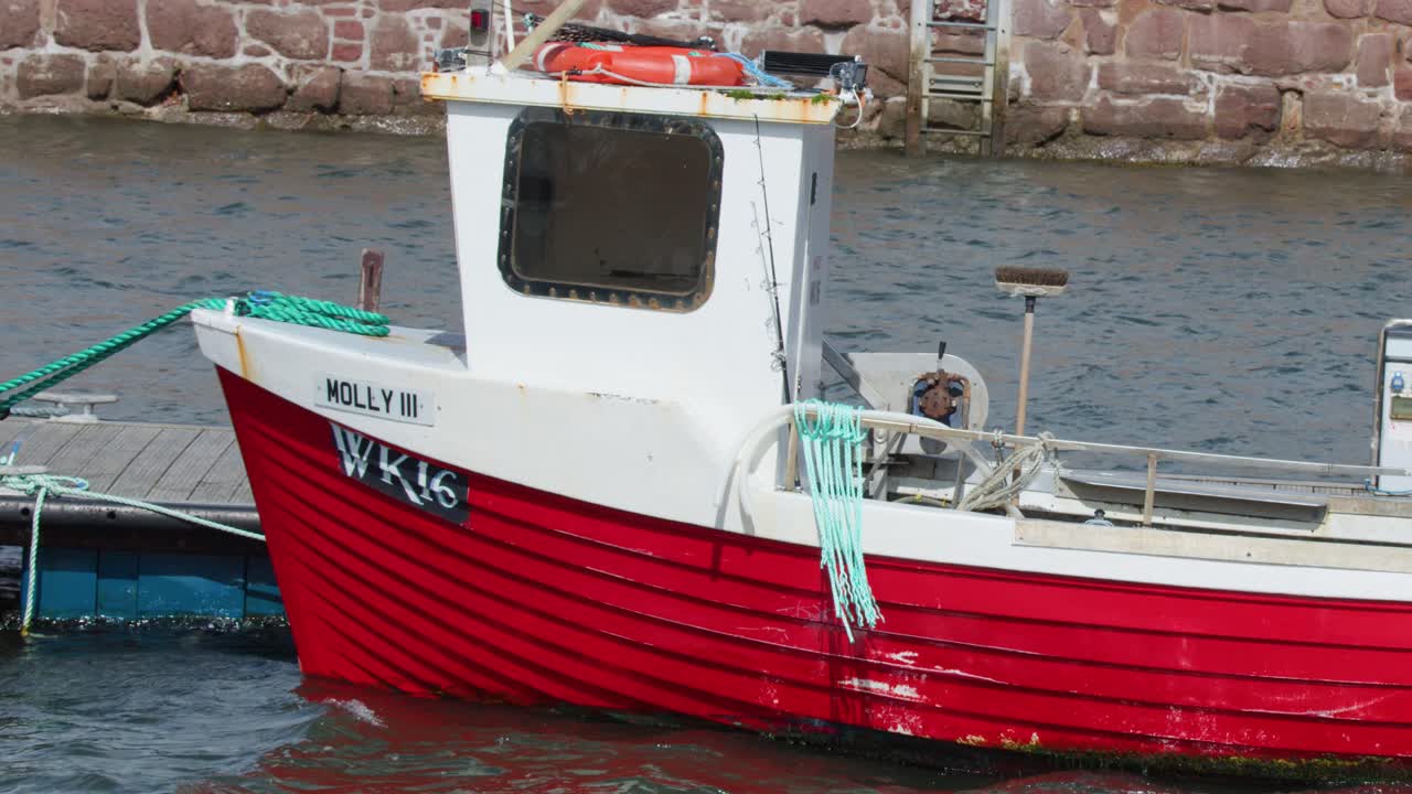Red fishing boat nears stone dock in daylight, gentle pan, calm harbor, overcast lighting