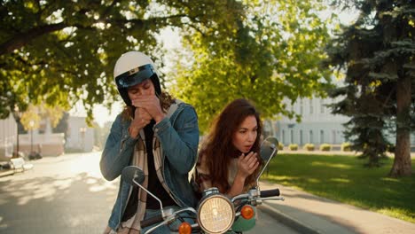 A-brunette-girl-preens-herself-looking-into-the-side-mirror-of-a-moped,-and-her-boyfriend-in-a-denim-shirt-in-a-White-helmet-pretentiously-smokes-a-cigarette-and-looks-at-his-girlfriend-in-a-sunny-summer-city-on-the-street