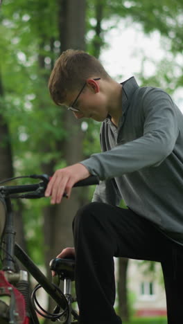 joven con gafas sostiene el manillar de una bicicleta estacionada en un campo cubierto de hierba, ajustando el manillar firmemente mientras comprueba la funcionalidad de los frenos, se detiene para ajustar sus zapatillas