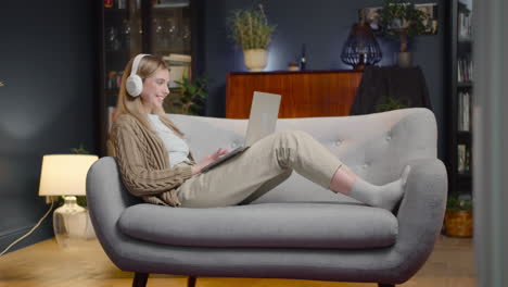 happy young woman with wireless headphones chatting on laptop computer while lying on couch at home