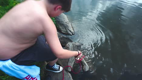 shirtless boy crouched by ariver, and pulling out magnet tied on long rope