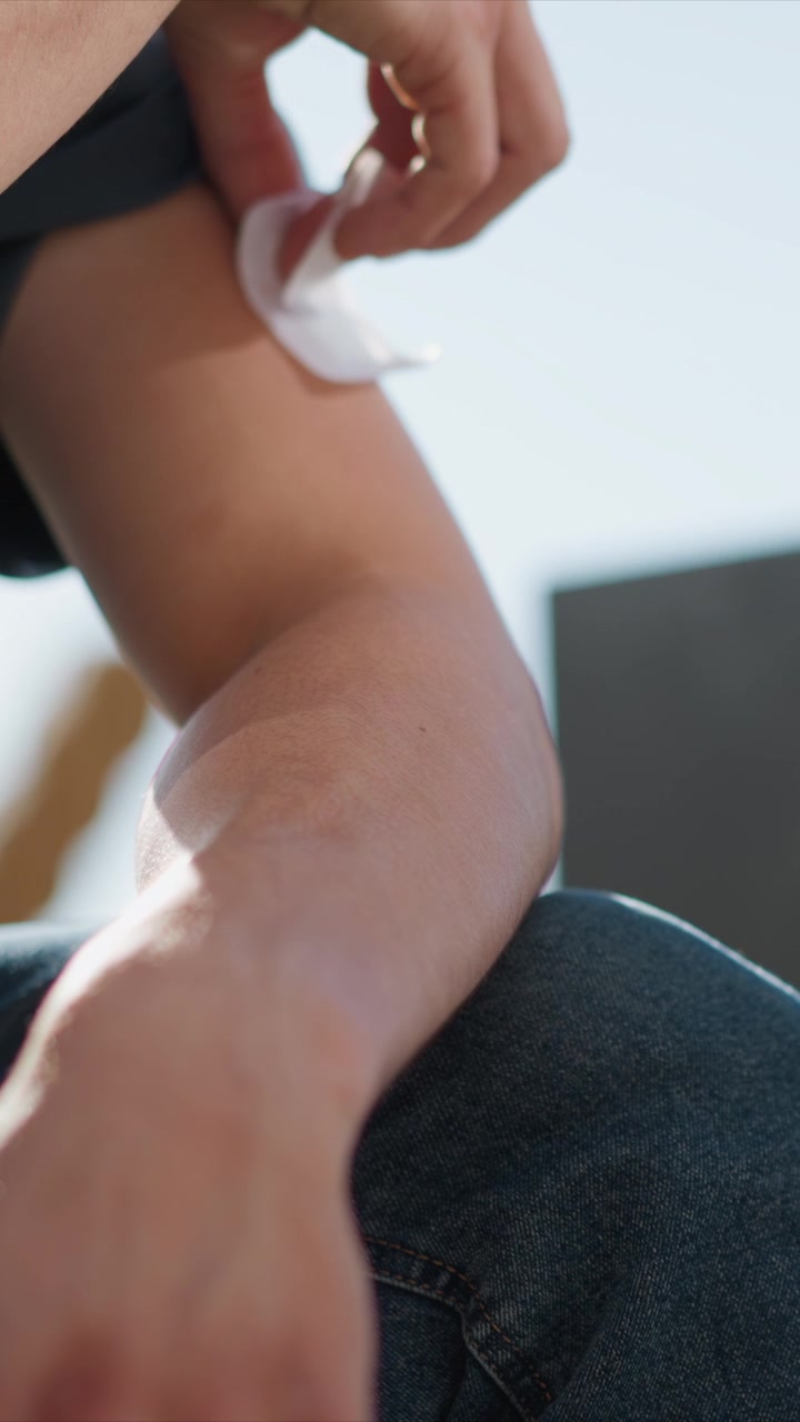 Primo piano di una persona che applica cerotti adesivi al braccio e alla mano, rivelando segni rossi sulla pelle. Il processo implica un posizionamento attento con un cuscinetto bianco visibile contro la pelle nuda, che enfatizza.