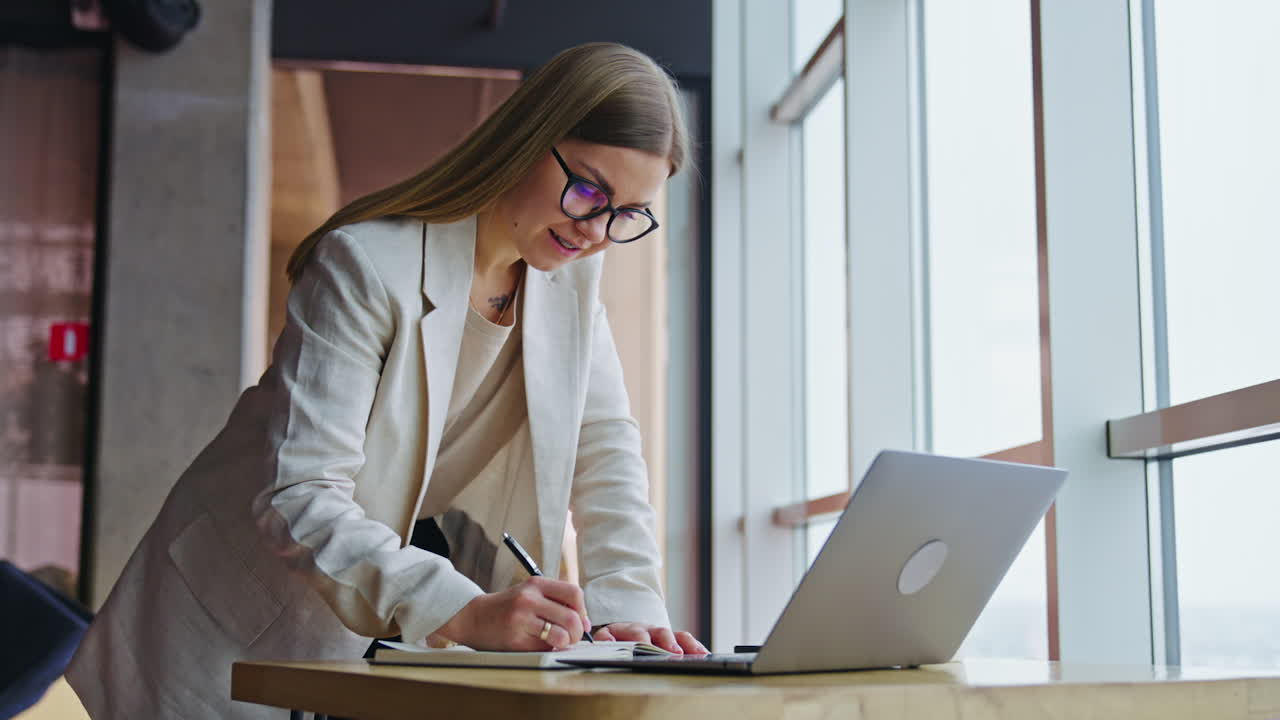 Office lady surfing the net and taking notes. Female employee standing leaned on the table in a spacious office.
