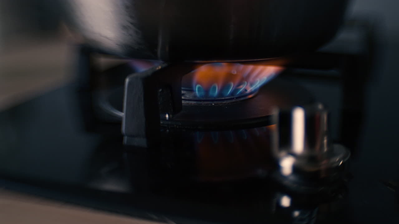 A distorted shot of a black ceramic gas stove hob being turned on or ignited, flames appear and an aluminium pot is put on the stove