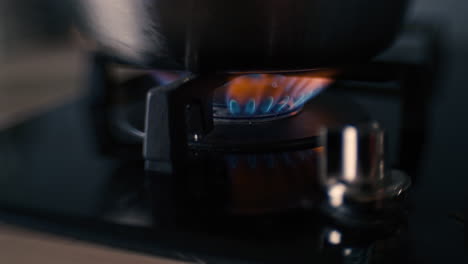 a distorted shot of a black ceramic gas stove hob being turned on or ignited, flames appear and an aluminium pot is put on the stove