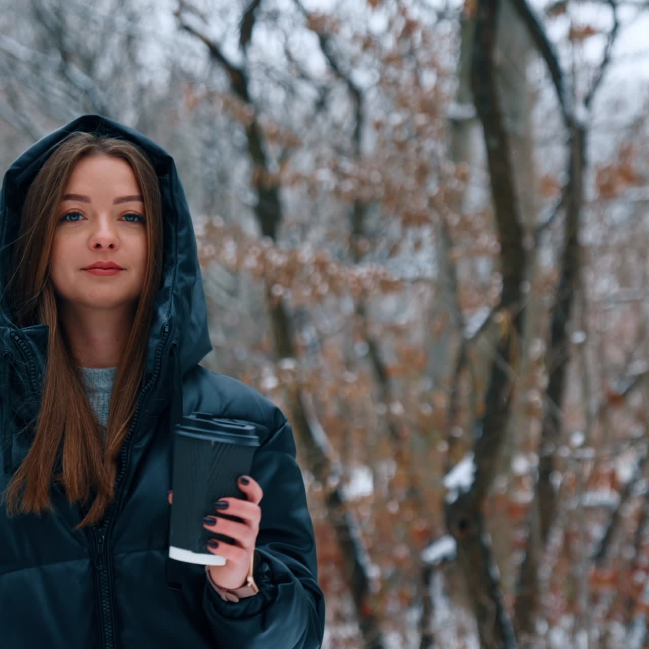 Donna caucasica con i capelli lunghi in blazer nero che cammina nel parco. La donna tiene in mano un bicchiere di carta e si gode la giornata invernale.