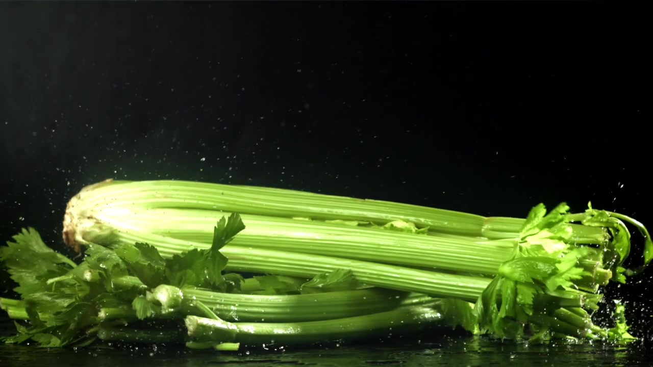 Gambi di sedano verde fresco che cadono e schizzano su una superficie nera bagnata