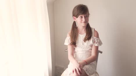 Caucasian-Girl-Child-Sitting-on-Chair-By-Window-in-White-Dress-and-Hair-in-Pigtails-Crossing-Her-Legs-and-Looks-With-Smile-in-Opposite-Direction-in-Room-and-Smiles-To-Somebody