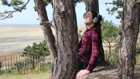 shot of an authentic hipster man listening to music with headphones whilst sitting in the sun against a tree trunk