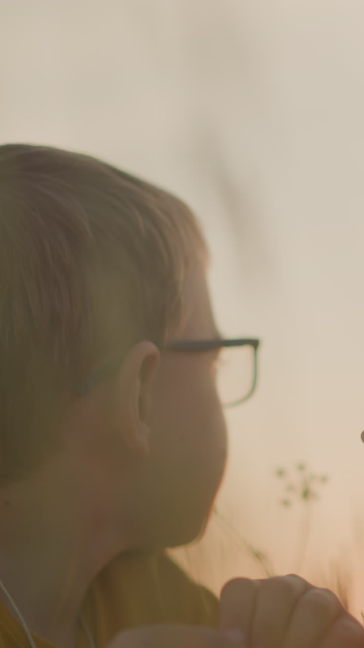 un momento tierno cuando una mujer con un vestido azul ofrece una flor púrpura a un niño con gafas, invitándolo a olerla durante una serena puesta de sol, capturando una conexión pacífica con la naturaleza