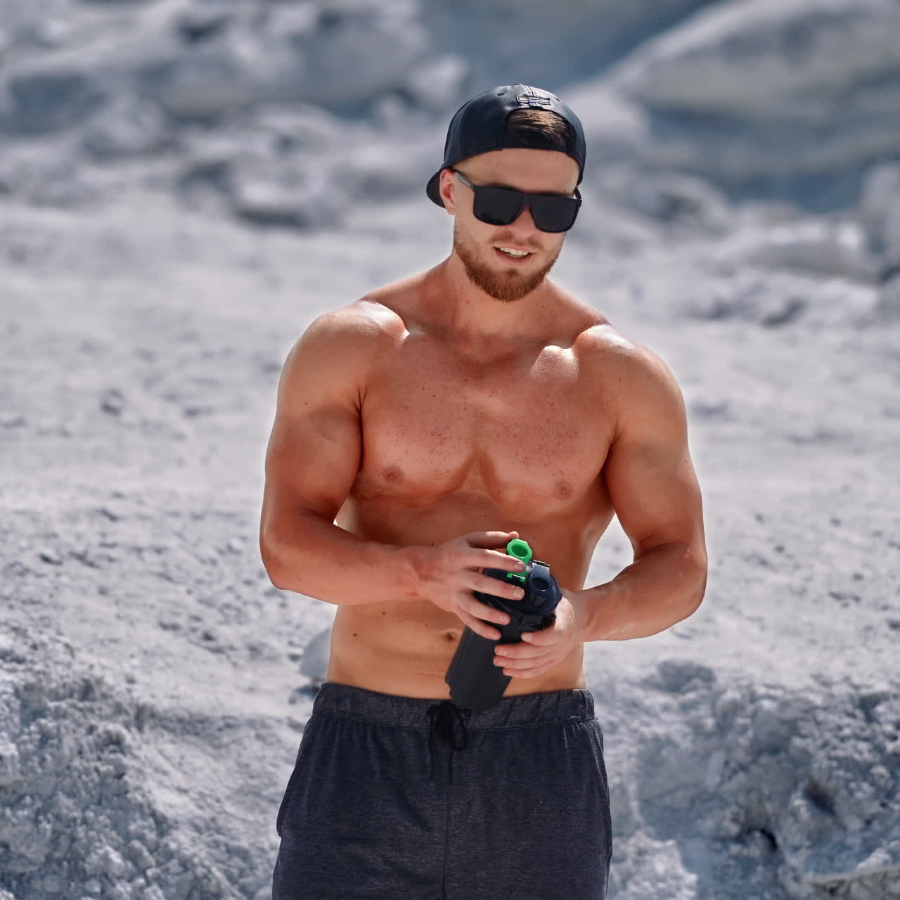 Bearded athlete with black flask in nature. Shirtless muscular bodybuilder drinking water on white rocky background in summertime.