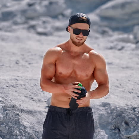 bearded athlete with black flask in nature. shirtless muscular bodybuilder drinking water on white rocky background in summertime.