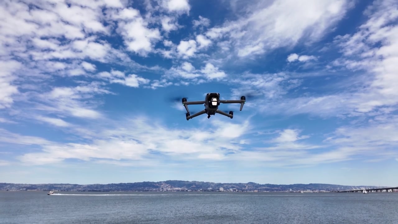 Drone consumer moderno che plana in un bellissimo cielo blu con nuvole sparse, in lenta ascesa da sopra uno specchio d'acqua con un ponte e uno skyline cittadino visibile in lontananza.