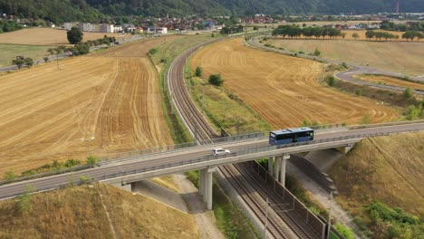 blue bus drives on the bridge over the rails in rural country side between yellow fields - aerial drone shot