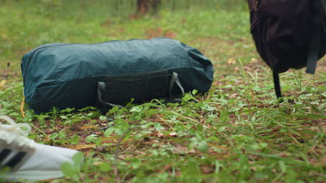 close up of campers dropping heavy duffel bag on forest floor, focus on green bag strap brushing ground, pine needles and small plants under it, conveying rest pause in woodland adventure
