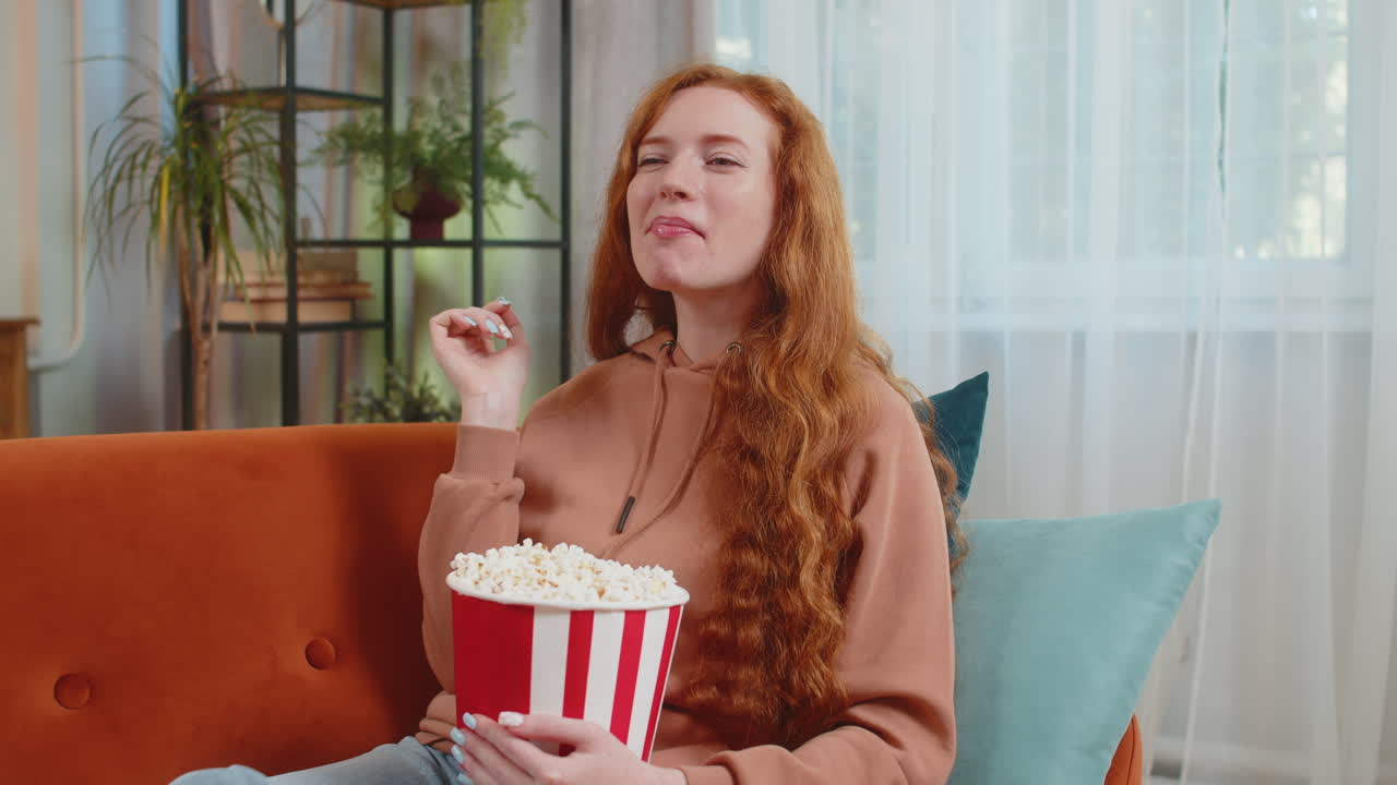 mujer pelirroja sonriente comiendo palomitas de maíz y viendo una película expresión wow sentada en el sofá en casa