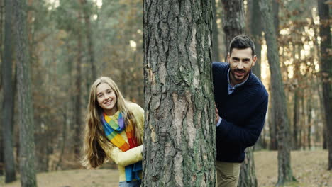 portrait shot of caucasian handsome dad and his cute little girl looking at camera behind a tree trunk in the forest