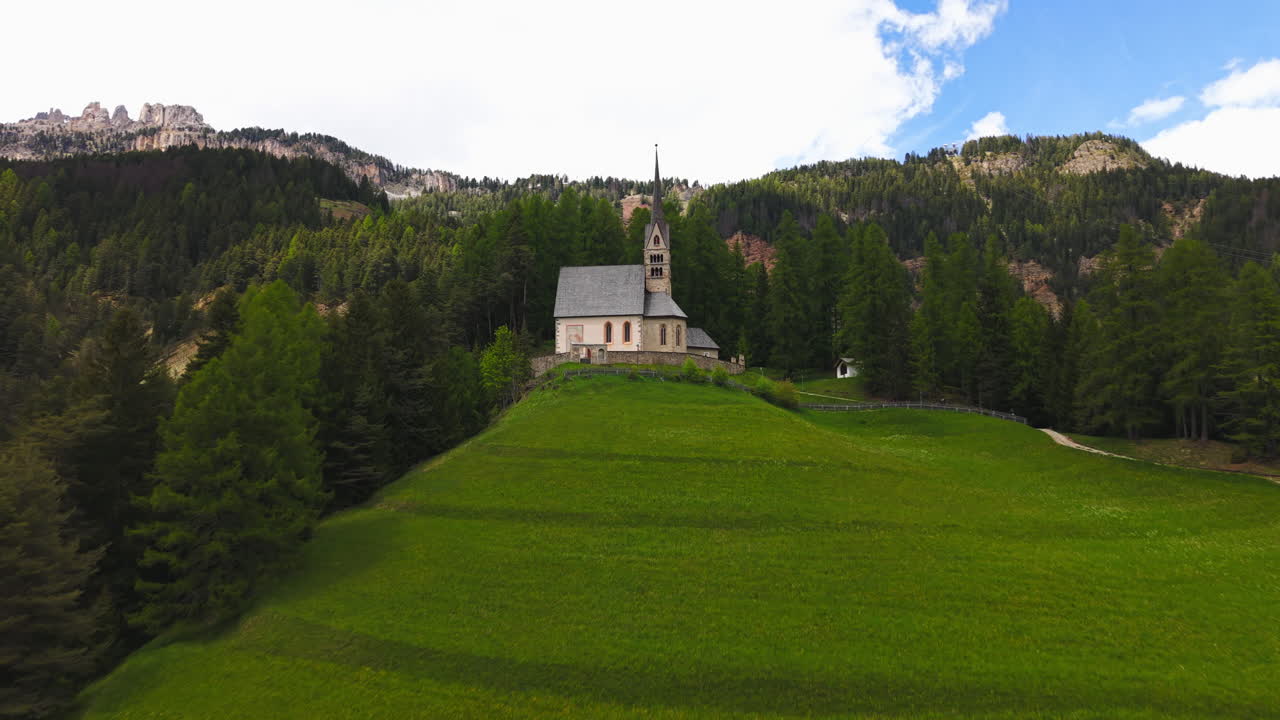 Drone footage of a historic alpine church surrounded by lush green meadows and forested mountains in the Dolomites, Italy