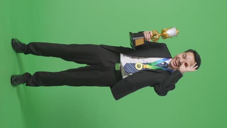 full body of asian business man in a suit and tie with a gold medal and trophy showing okay hand sign over eye and smiling to camera as the first winner on green screen background in the studio