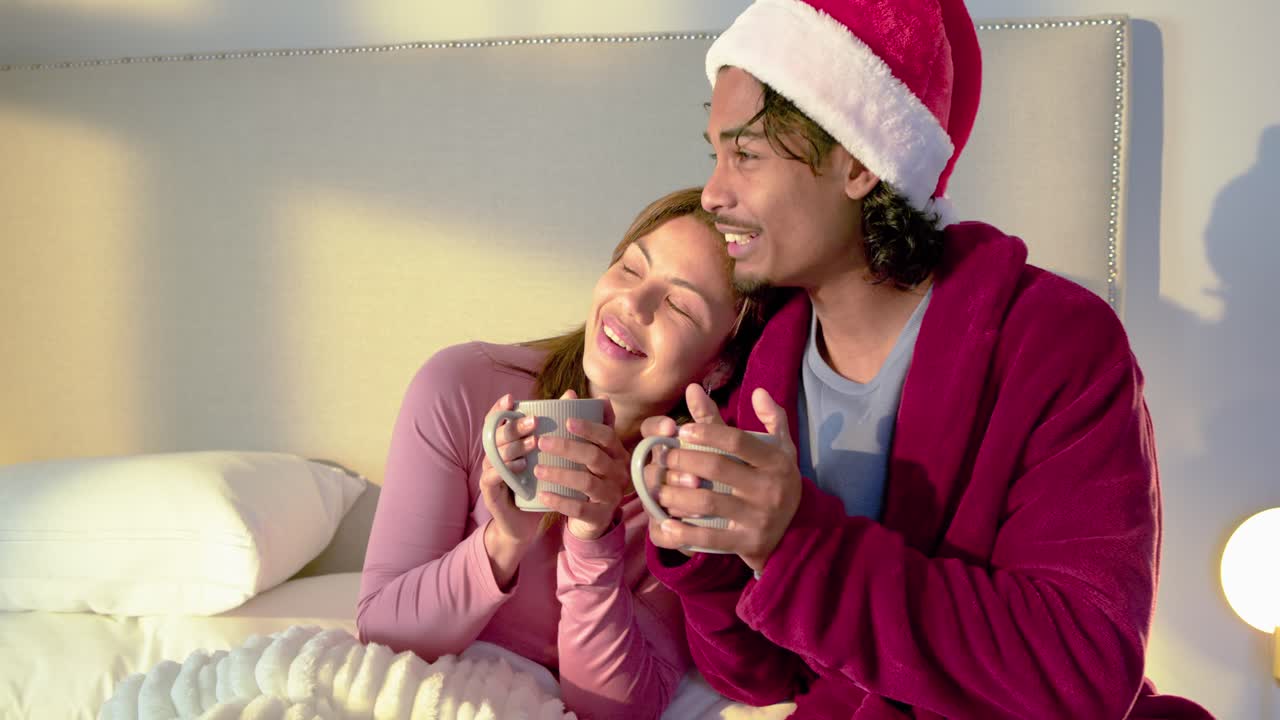 Couple on bed under grid light, man wearing Santa hat smiling and sparking chatter with mug-sipping
