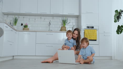tecnología moderna. apartamento moderno amorosa mamá y dos hijos pequeños sentados en el suelo en la sala de estar miran la pantalla de la computadora portátil. niños con mamá juegan en una computadora portátil.