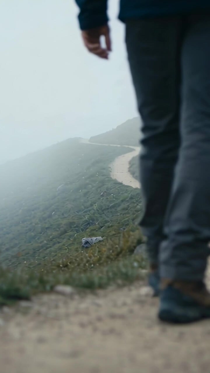 Vertical video: Low shot starting, man in dark coat walking hill path with boots, mist, copy space