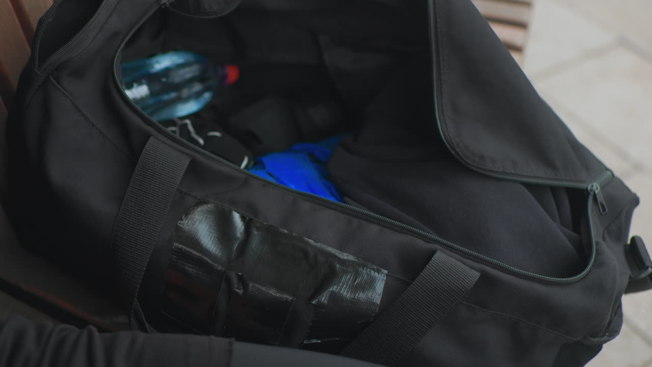 Una persona bianca scopre una custodia blu all'interno di una sacca da viaggio, le dita tracciano la cucitura nastrata ed estraggono un organizer luminoso. Primo piano di avambracci tatuati su un pavimento piastrellato, sorpresa tattile e ispezione accurata.