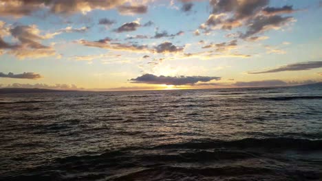 Vista-De-La-Playa-Rocosa-De-Las-Rocas-Que-Se-Elevan-Más-Allá-De-Las-Olas-Hasta-La-Puesta-De-Sol-En-El-Fondo-Con-Nubes-Encima