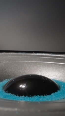 speaker rotates in circle and plays music. blue powder bounces lying in central part of diaphragm. modern portable music player works in room with gray walls macro