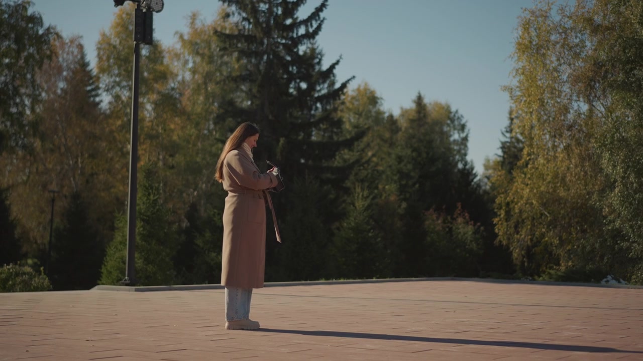 Primo piano di una donna in piedi all'aperto in autunno, che tiene in mano e guarda il suo smartphone. La donna indossa un lungo cappotto beige, jeans azzurri e scarpe bianche, contro un'area pavimentata aperta con alberi alti