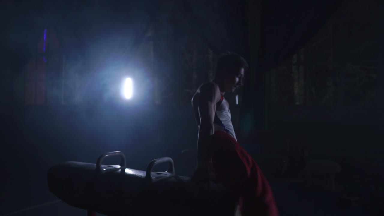 gimnasta masculino realiza ejercicios del programa olímpico en un caballo de pommel realizando giros y rodajes en cámara lenta preparándose para los juegos olímpicos entrenando duro en la habitación oscura alrededor del humo y la luz del mostrador