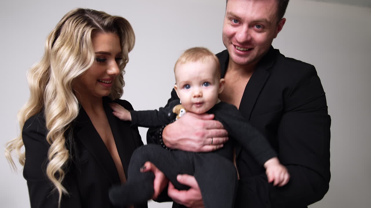 Caucasian parents wearing black jackets play with their little baby. Happy couple entertain their child. White backdrop.