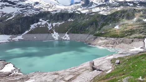 Aguas-Tranquilas-De-Un-Embalse-En-La-Parte-Inferior-De-Montañas-Cubiertas-De-Nieve,-Antena