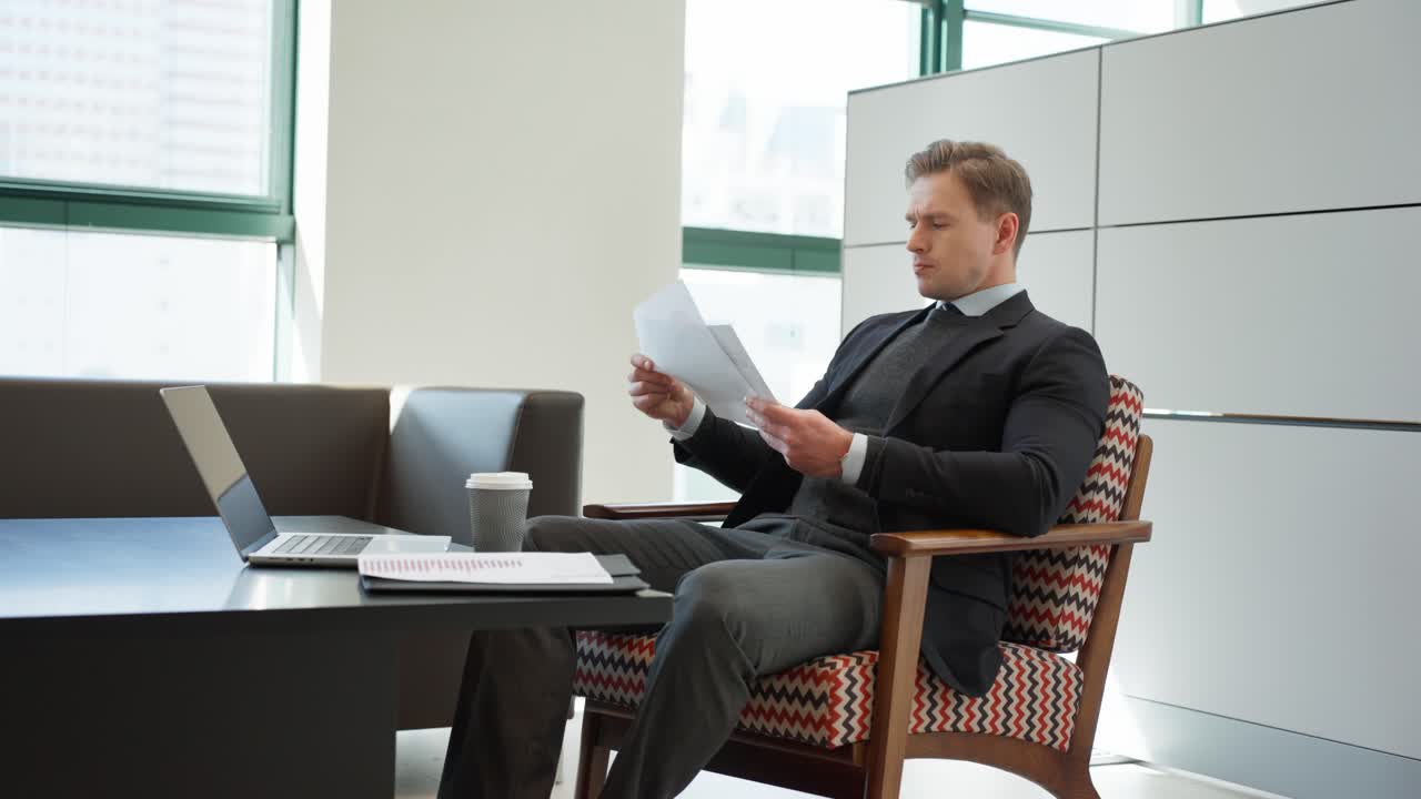 Un uomo d'affari in abito scuro esamina documenti mentre è seduto a una moderna scrivania d'ufficio attrezzata con un laptop e una tazza di caffè, circondato da pareti grigie minimaliste e grandi finestre - ripresa in parallasse