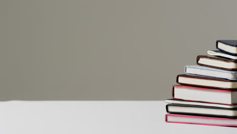 A-stack-of-books-with-various-covers-sits-on-a-white-surface-against-a-grey-background