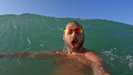 active bearded man with yellow sunglasses having fun splashing with big sea waves breaking on his head