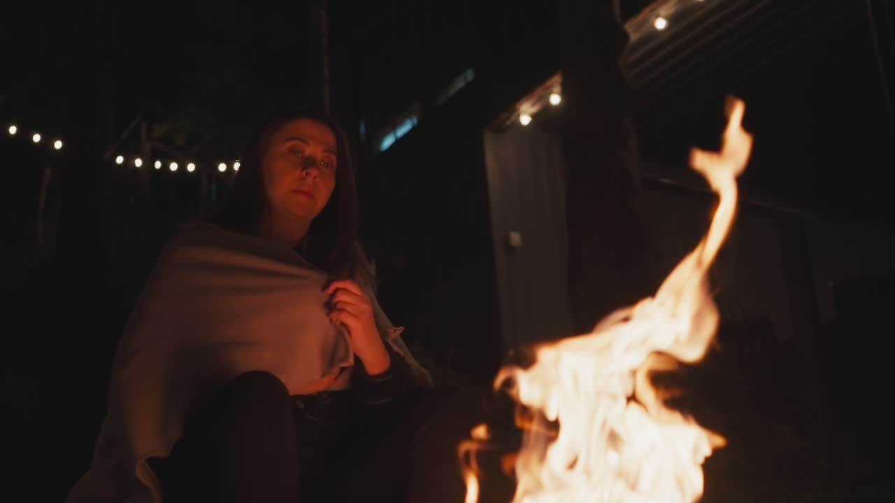 mujer aleja las manos de la llama del fuego. mujer solitaria se sienta frente a la hoguera con una manta sobre los hombros. señora calienta los dedos debido al clima frío