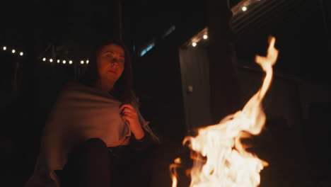 woman takes hands away from fire flame. lonely woman sits in front of bonfire with blanket over shoulders. lady warms fingers because of cold weather