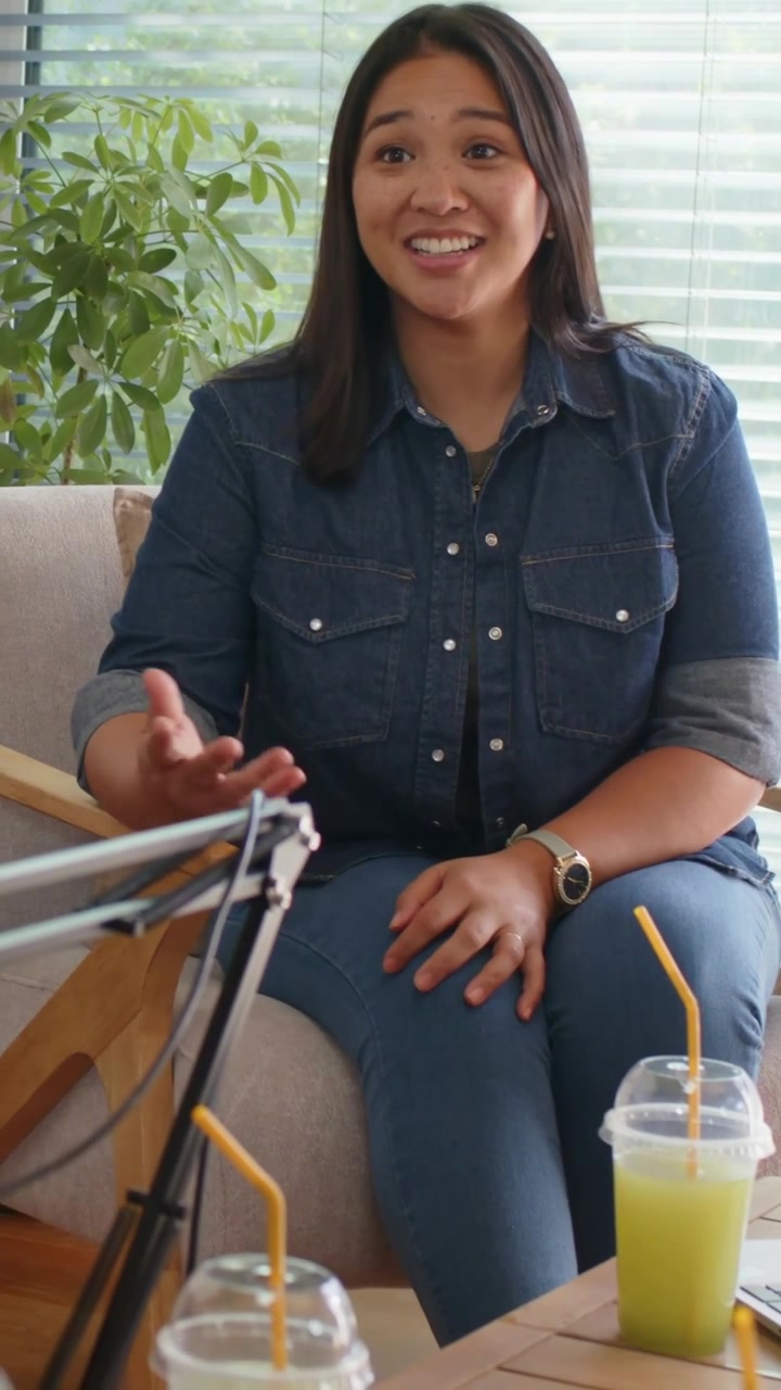 Vertical video: Woman in denim answering offscreen cue, gesturing at home with mic laptop cups