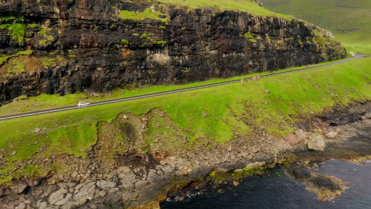 A winding road hugs a green hillside with cliffs in the Faroe Islands by the coast