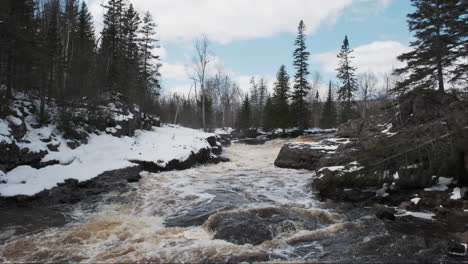 Rocky-winter-river-carving-its-path-beneath-a-blue-sky-with-wispy-clouds-and-a-towering-forest-of-pines