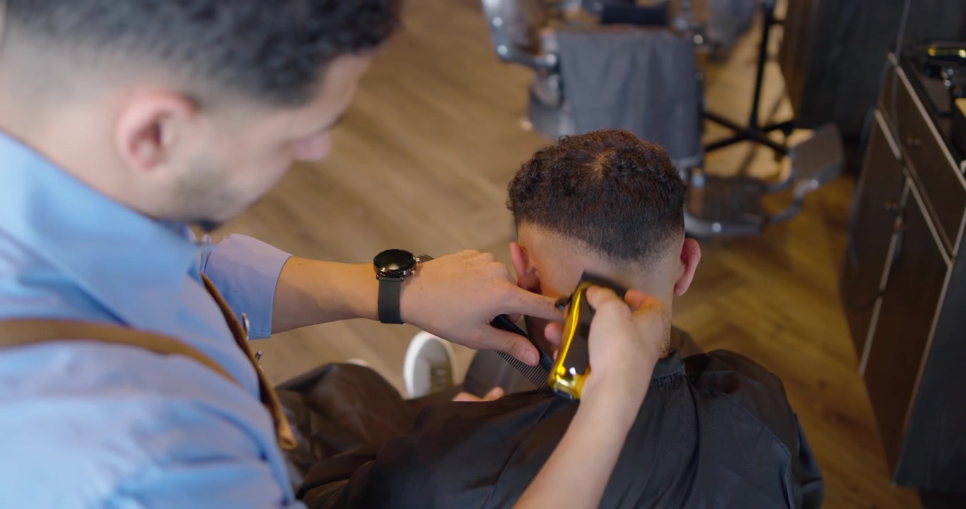 Barbiere afroamericano che taglia i capelli a un cliente in un negozio, creando una sfumatura con un tagliacapelli dorato, dopo avergli inclinato la testa.