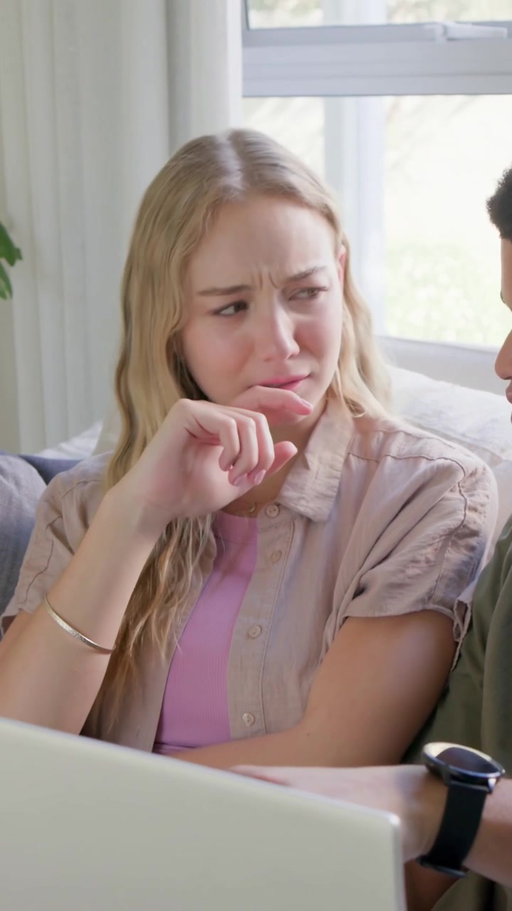 Vertical video: Glancing at laptop adult woman in button-up tapping chin on couch, weighing options