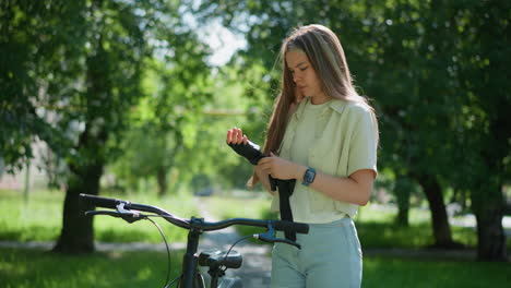 young woman in jeans short and smart watch stands beside her bicycle, focused on removing her biker glove under gentle sunlight filtering through leafy trees