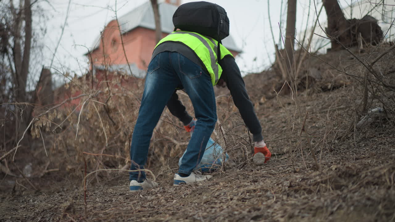 Volunteer in reflective vest and hoodie carrying backpack and plastic bag filled with collected litter during environmental cleanup in dry overgrown area near historic brick building