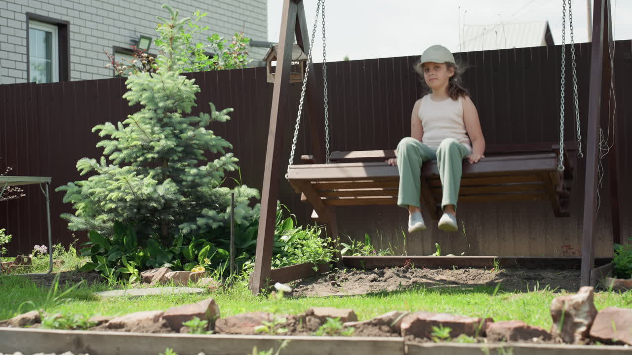 White Girl Approaches Wooden Garden Swing With Evergreen Tree Nearby, Summer Sunlight, Casual Cap And Green Pants, Hesitant Steps, Quiet Backyard Mood, Hands Touching Bench And Observing Surroundings