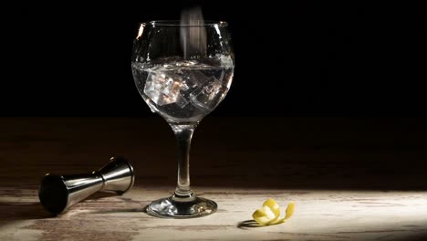 Person-pouring-ice-cubes-into-glass-of-gin-tonic-with-lemon-and-jigger-on-table-in-dark-room