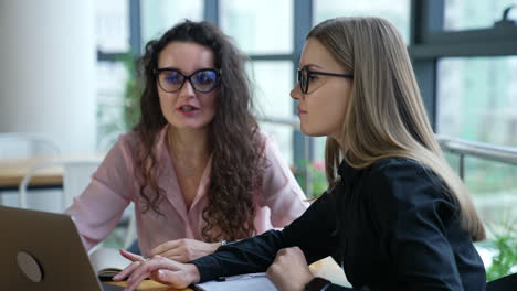 women coworkers discuss working moments at the table. female employees look at laptop and talk. blurred office background.