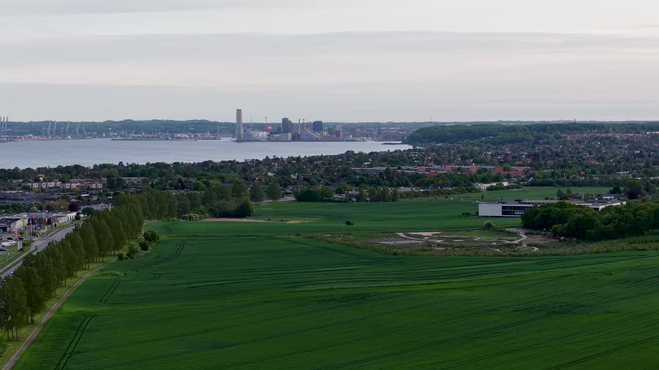 Aerial drone view of green farmland leading into a Danish city with distant coastline and cloudy sky